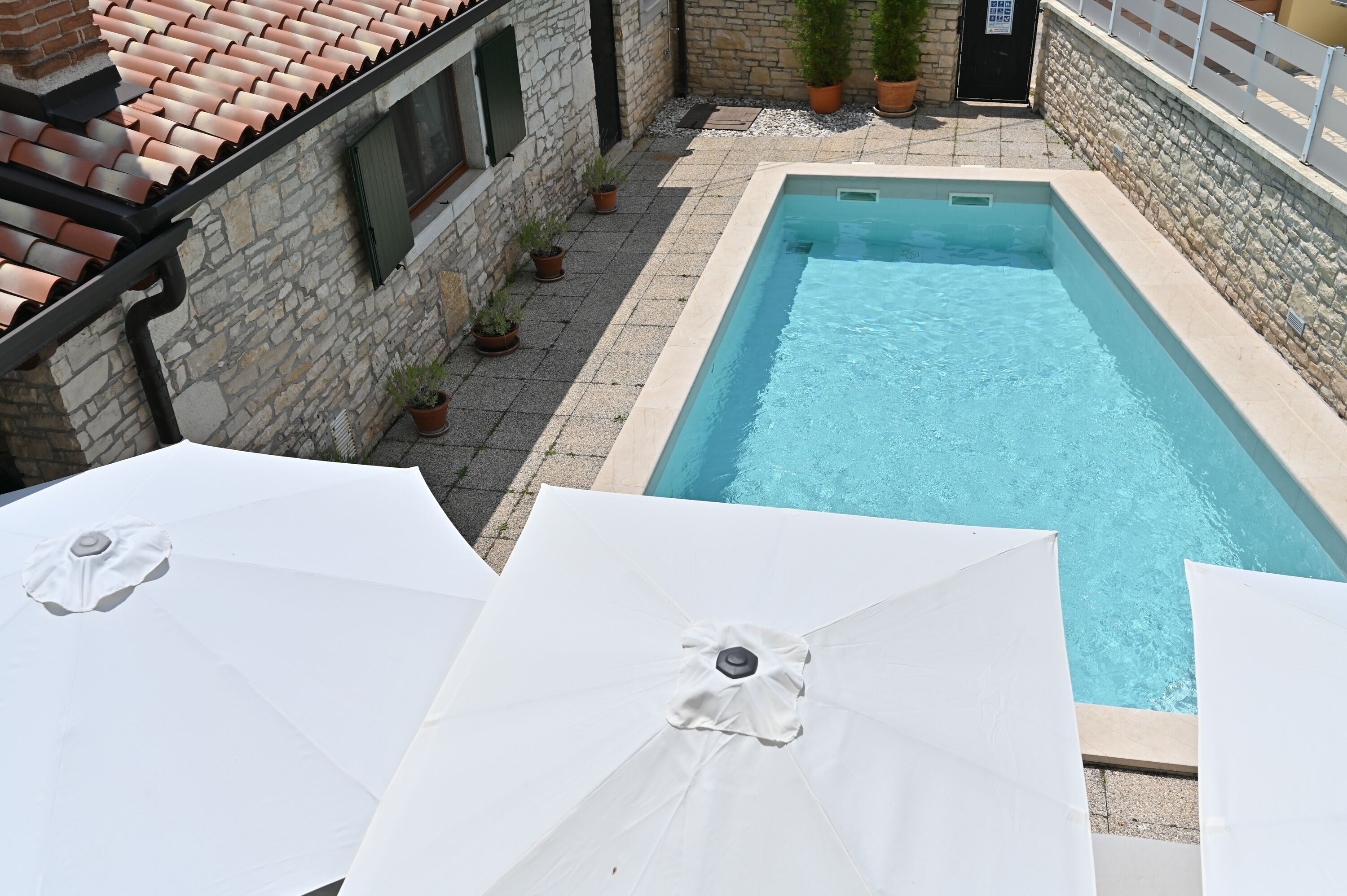 A swimming pool with a stone wall and tiled roof, surrounded by potted plants and umbrellas.