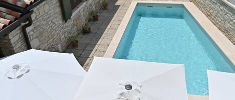 A swimming pool with a stone wall and tiled roof, surrounded by potted plants and umbrellas.