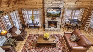 A cozy living room with a stone fireplace, a television mounted above it, and a dining area with a table and chairs. - Spacious 4BR Cabin w/ Game Room, Hot Tub & Pools – 3 Miles to Parkway (Sevierville)