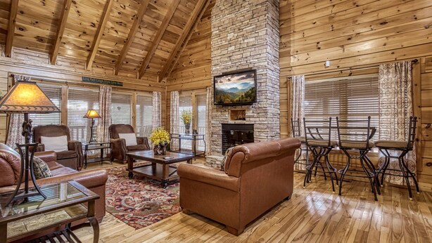 A cozy log cabin interior with a stone fireplace, leather furniture, and a large TV mounted on the wall. - Spacious 4BR Cabin w/ Game Room, Hot Tub & Pools – 3 Miles to Parkway (Sevierville)