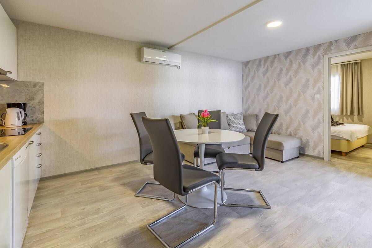 Cozinha moderna com cafeteira, sala de jantar com mesa redonda e cadeiras, sofá e uma cama no quarto vizinho.