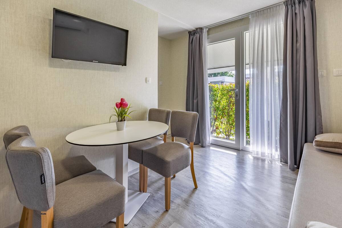 Quarto moderno com TV de tela plana na parede, uma mesa de jantar redonda com quatro cadeiras, uma pequena planta em vaso e um sofá.