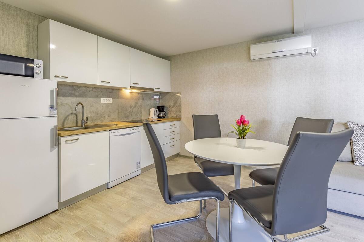 Cozinha compacta com armários brancos, micro-ondas, máquina de café e lava-louças. Há uma mesa redonda com quatro cadeiras e um vaso de planta.