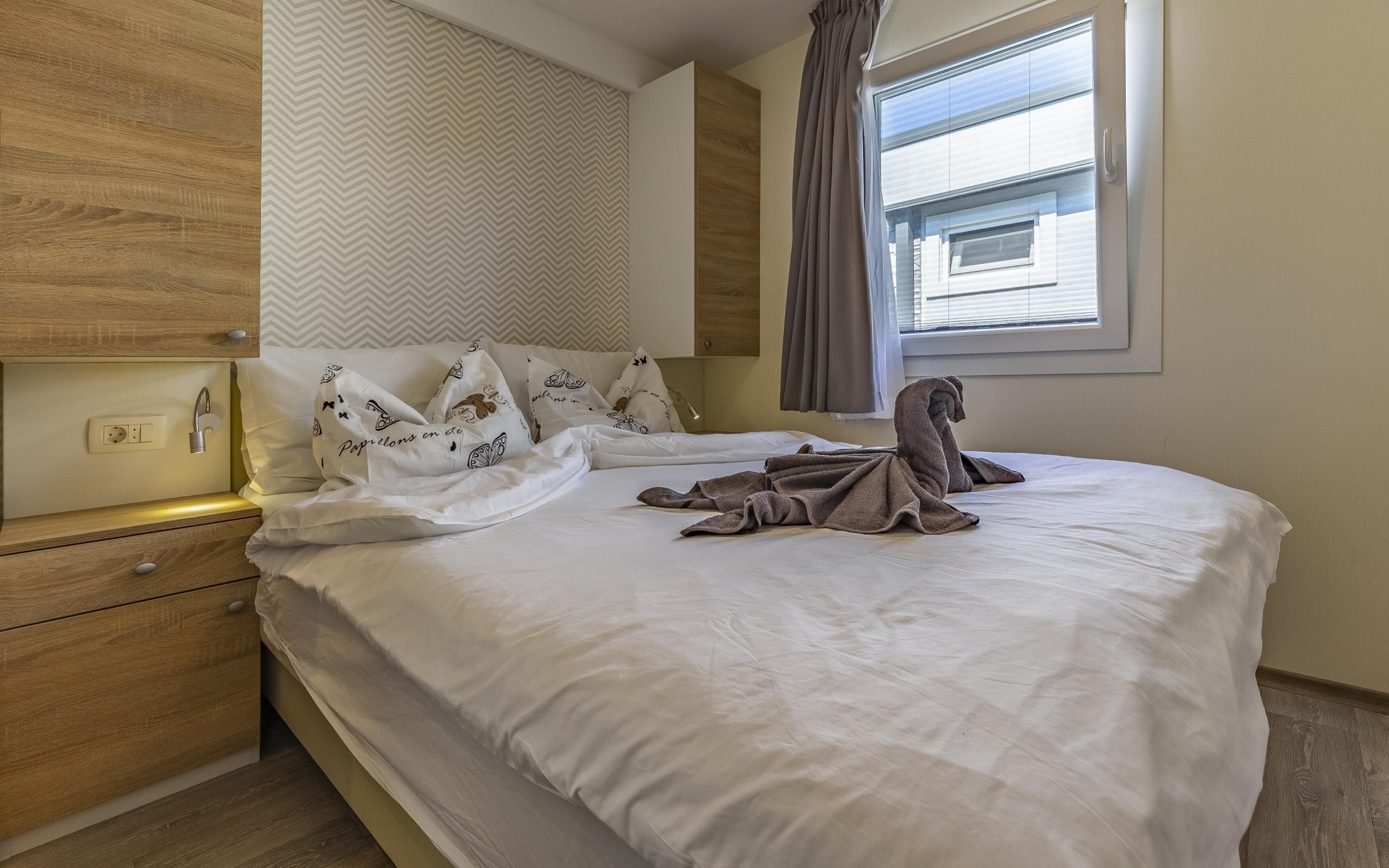 A neatly made bed with white linens and a towel, a wooden nightstand with a lamp, and a window with blinds.