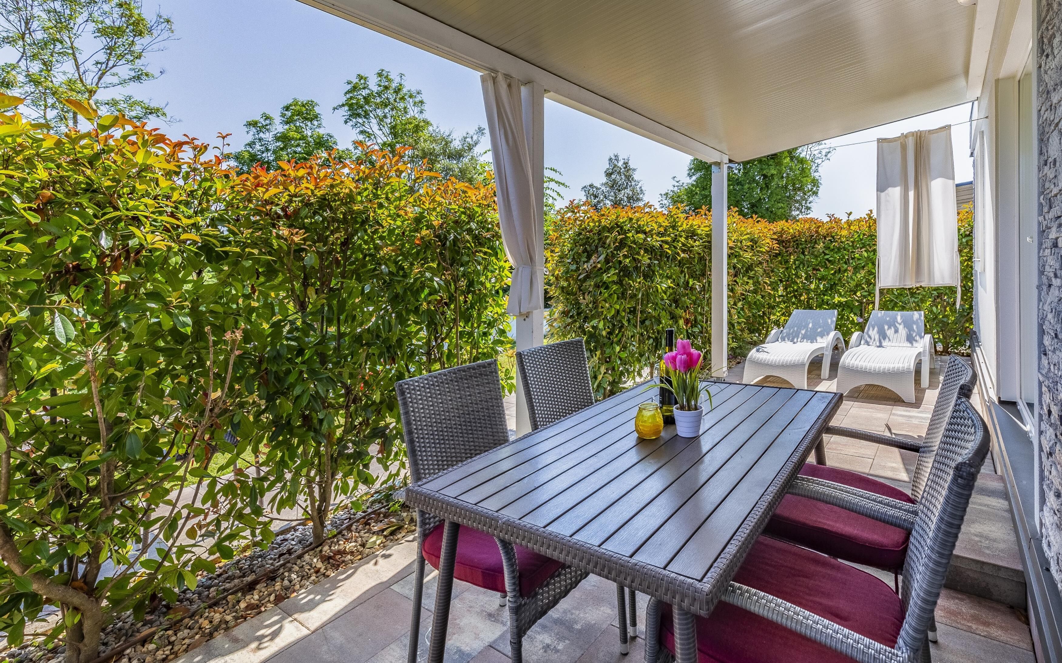 Une terrasse couverte, aménagée avec des meubles en osier et une table, le tout entouré d’une végétation luxuriante.