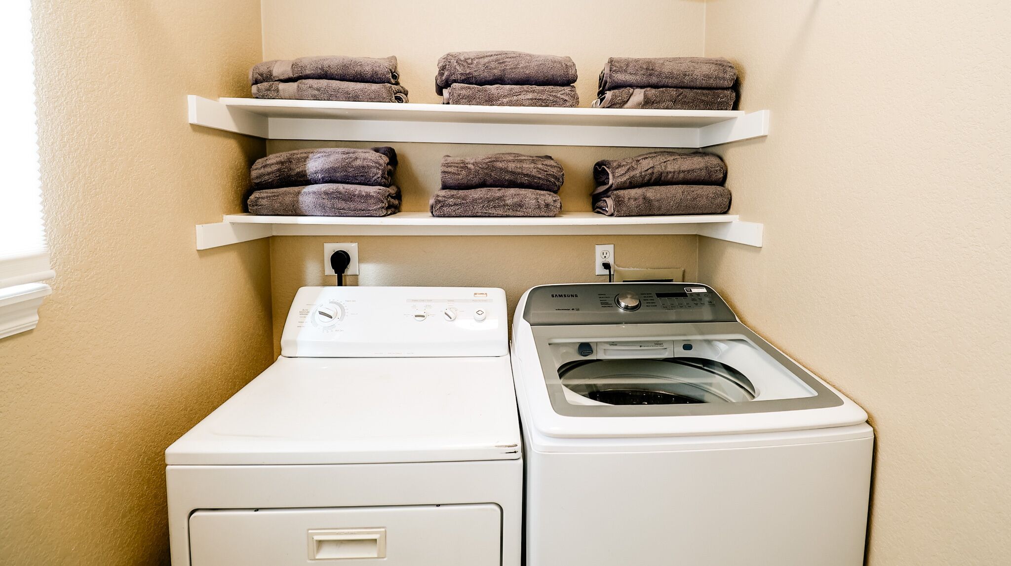 A laundry area with a front-loading washer and dryer, two shelves above holding folded towels, and a power outlet on the wall.
