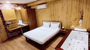 Habitación de hotel con piso de madera, una cama grande con ropa blanca, cabecera de madera, tocador de madera con espejo y bañera blanca con zona de ducha.