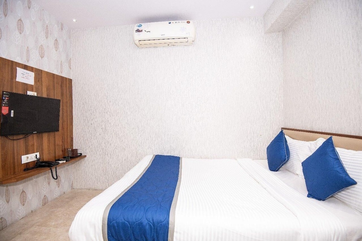 A hotel room with a bed, blue pillows, a TV, and an air conditioning unit.