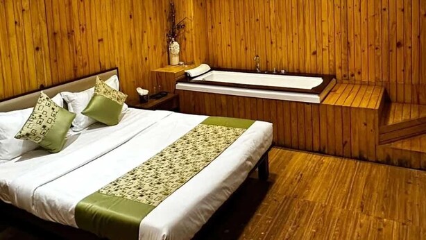 Una habitación con un muro revestido de paneles de madera, una cama con ropa blanca y detalles verdes, una mesita de noche con un florero, y una bañera.