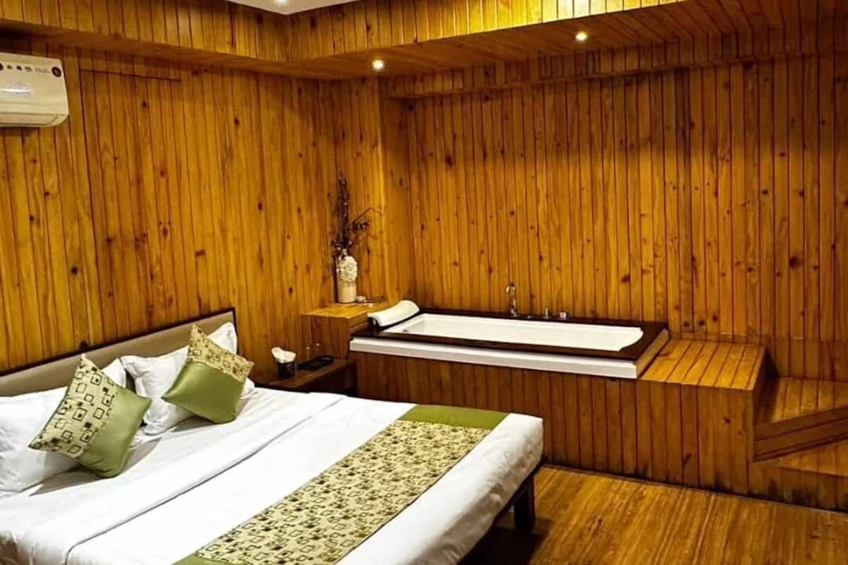 A room with a wooden paneled wall, a bed with white bedding and green accents, a small bedside table with a vase, and a bathtub.