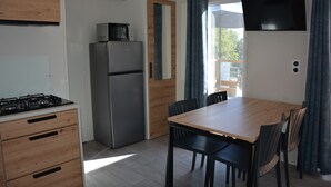 A compact kitchen with a gas stove, a refrigerator, and a wooden dining table with chairs. - Bungalow Neuf Superbe Terrasse Plein sud Face mer à l' ile d' or Camping 4 (Saint-Raphaël)