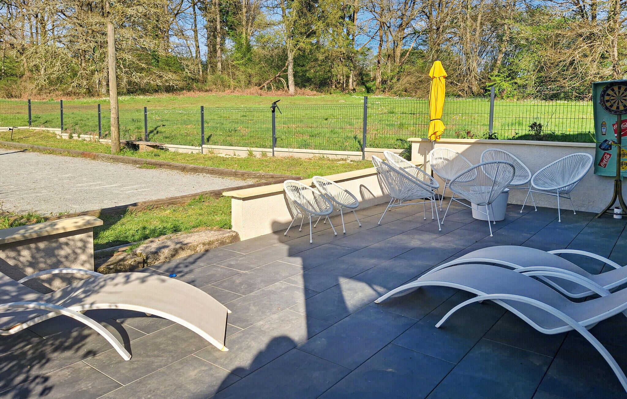 Een terras met witte stoelen en een gele parasol, omringd door een groene hekken en bomen.