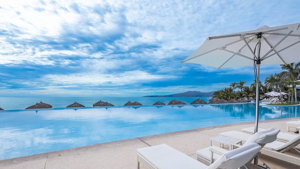 A pool with lounge chairs and umbrellas, overlooking a calm sea with palm trees and thatched huts. - NEW Beach Club Game Room Cinema Condos (Nuevo Nayarit)