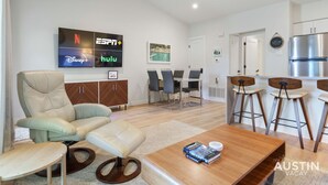Un salon moderne comprenant un canapé, une table basse, un téléviseur fixé au mur diffusant des services de streaming, un coin repas avec une table et des chaises, et un coin cuisine avec un réfrigérateur et des tabourets de bar.