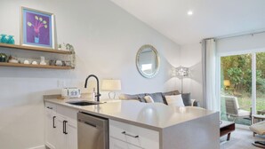 Une cuisine moderne dotée d’un îlot central, de meubles encastrés et d’un lave-vaisselle. On y trouve également un coin repas avec un canapé et des fauteuils, un grand miroir et une fenêtre donnant sur un espace verdoyant.