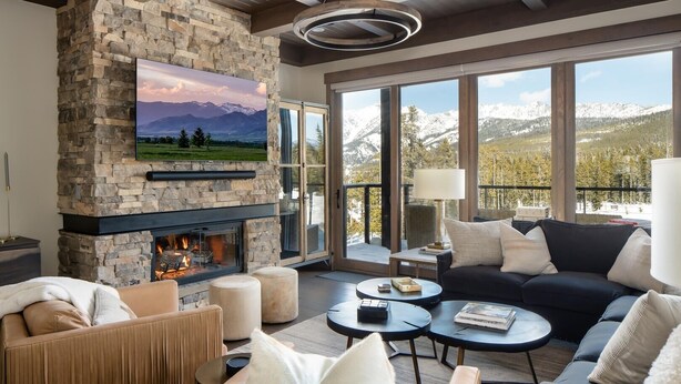 Ein modernes Wohnzimmer mit Kamin aus Stein, gemütlicher Sitzgruppe, Fernseher und großen Fenstern mit Blick auf die Berge.