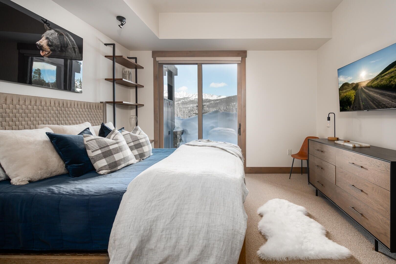 A modern bedroom with a large bed, a mounted bear head, a TV, and a view of a snowy landscape through a sliding door.
