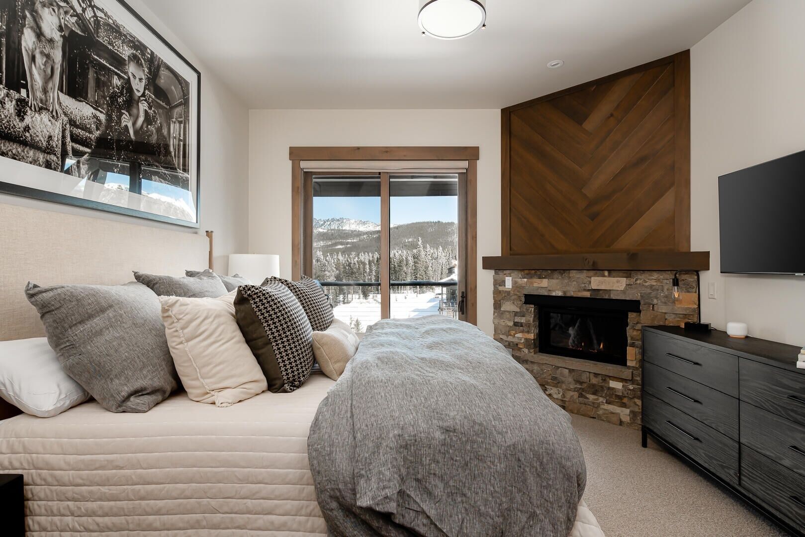 A bedroom with a large bed, a fireplace, a TV, and a view of a snowy landscape.