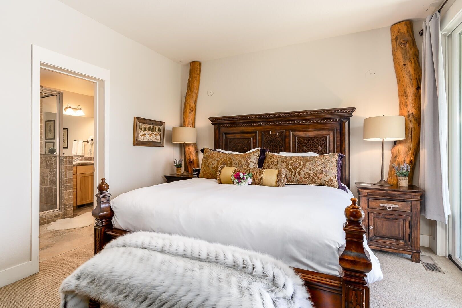 A spacious bedroom with a large bed, wooden headboard, two bedside tables with lamps, and a view of the bathroom.