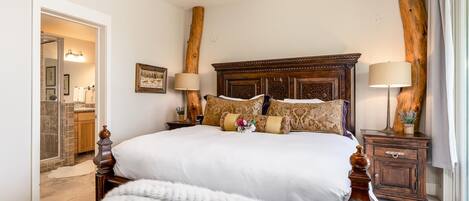 A spacious bedroom with a large bed, wooden headboard, two bedside tables with lamps, and a view of the bathroom.