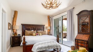 A bedroom with a large bed, wooden headboard, two bedside tables with lamps, a chandelier, a wooden cabinet, and a view through a glass door. - Saddle Ridge V2 3 Bedroom Condo by Moonlight Basin Lodging (Big Sky)