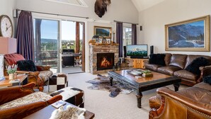 A spacious living room with a stone fireplace, leather furniture, and a mounted moose head. - Saddle Ridge V2 3 Bedroom Condo by Moonlight Basin Lodging (Big Sky)