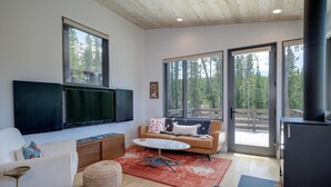 A modern living room with a sofa, coffee table, and TV. There are large windows and a sliding door to the outside.