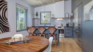 A modern kitchen with wooden chairs, a dining table with a fruit tray, and a large window.