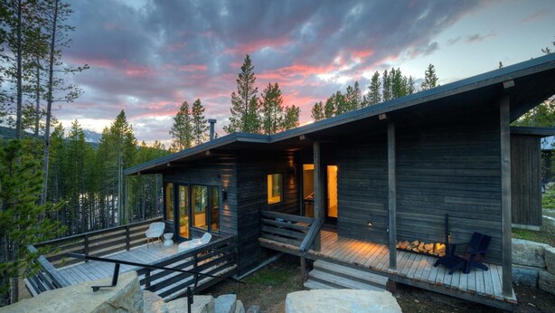 A modern cabin with a wooden exterior, a covered deck, and a view of the surrounding forest.