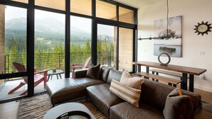 Ein modernes Wohnzimmer mit großem Fenster und Blick auf eine gebirgige Landschaft, einer Ledersofa, einem runden Couchtisch und einem Essbereich mit Holztisch und Bänken.