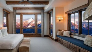 Een slaapkamer met een groot raam dat uitzicht biedt op de bergen, een bed met veel kussens, een bankje met kussens en een wandlamp.