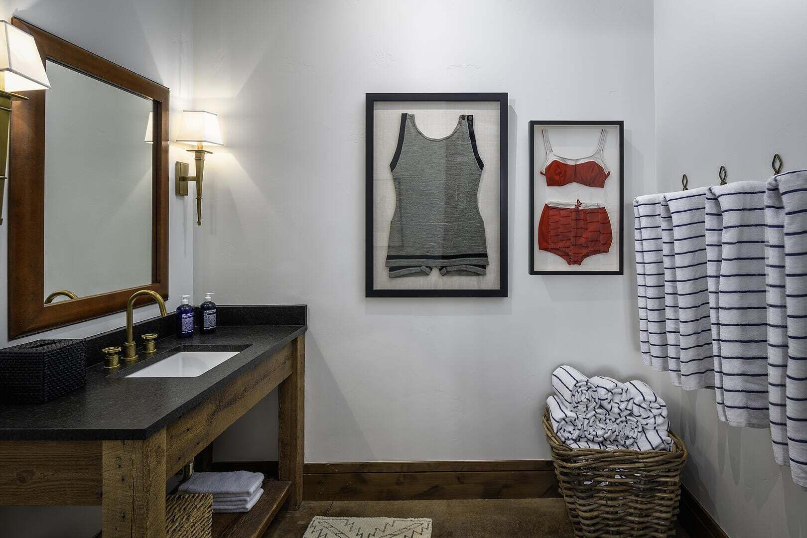 A bathroom with a wooden vanity, a large mirror, framed clothing on the wall, and a basket of folded towels.