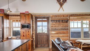 A rustic cabin interior with a wooden door, a kitchen area with a microwave, a bar counter, and a leather sofa.