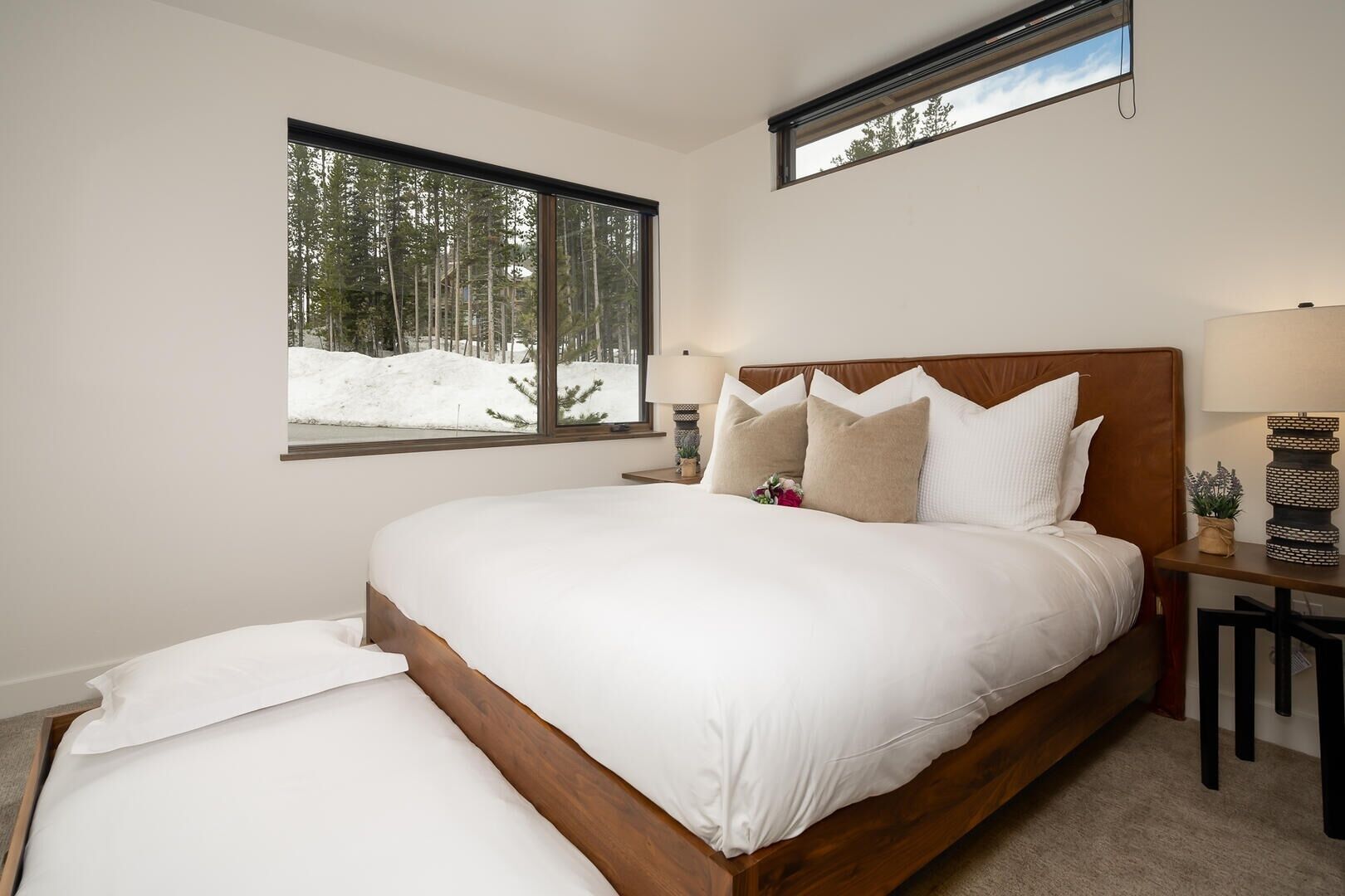 A bedroom with a large bed, a window with a view of trees and snow, a bedside table with a lamp, and a small plant on the nightstand.