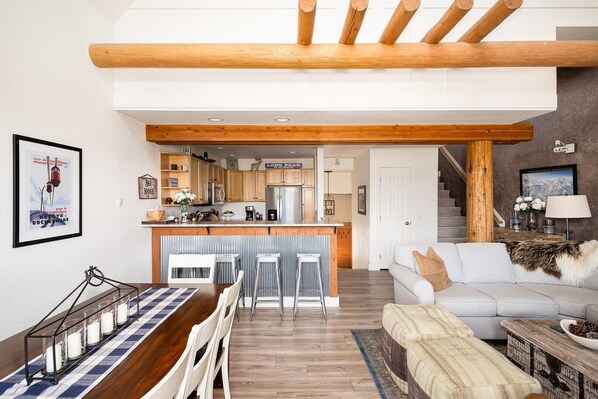 A modern kitchen with wooden beams, a dining area with a striped tablecloth, and a living room with a sofa and a coffee table.