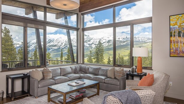 A spacious living room with a large sectional sofa, a coffee table, and a view of snow-capped mountains through large windows. - 18 Alpine Bend 6 Bedroom Home by Moonlight Basin Lodging (Big Sky)