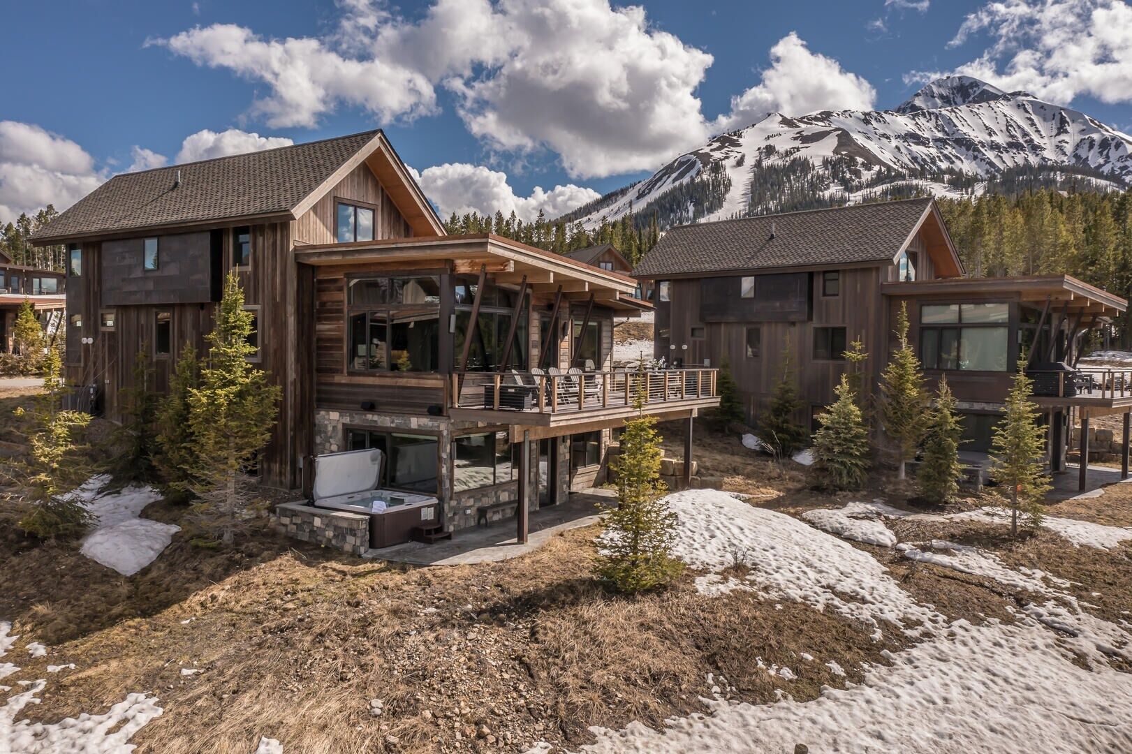 Una cabaña rústica moderna con terraza y jacuzzi, rodeada de nieve y árboles, con una montaña al fondo.