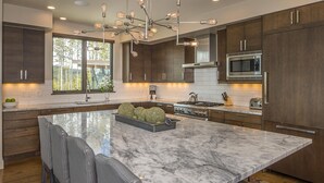 A modern kitchen with dark wood cabinets, a large marble island, and a central light fixture. - 18 Alpine Bend 6 Bedroom Home by Moonlight Basin Lodging (Big Sky)