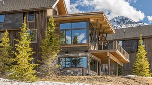 A modern log cabin with large windows, a stone foundation, and a view of a mountain range. - 18 Alpine Bend 6 Bedroom Home by Moonlight Basin Lodging (Big Sky)