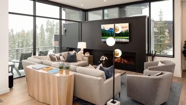 Un salon moderne avec un grand canapé, une table basse et une cheminée. Un téléviseur est fixé au mur et une fenêtre offre une vue sur les arbres.