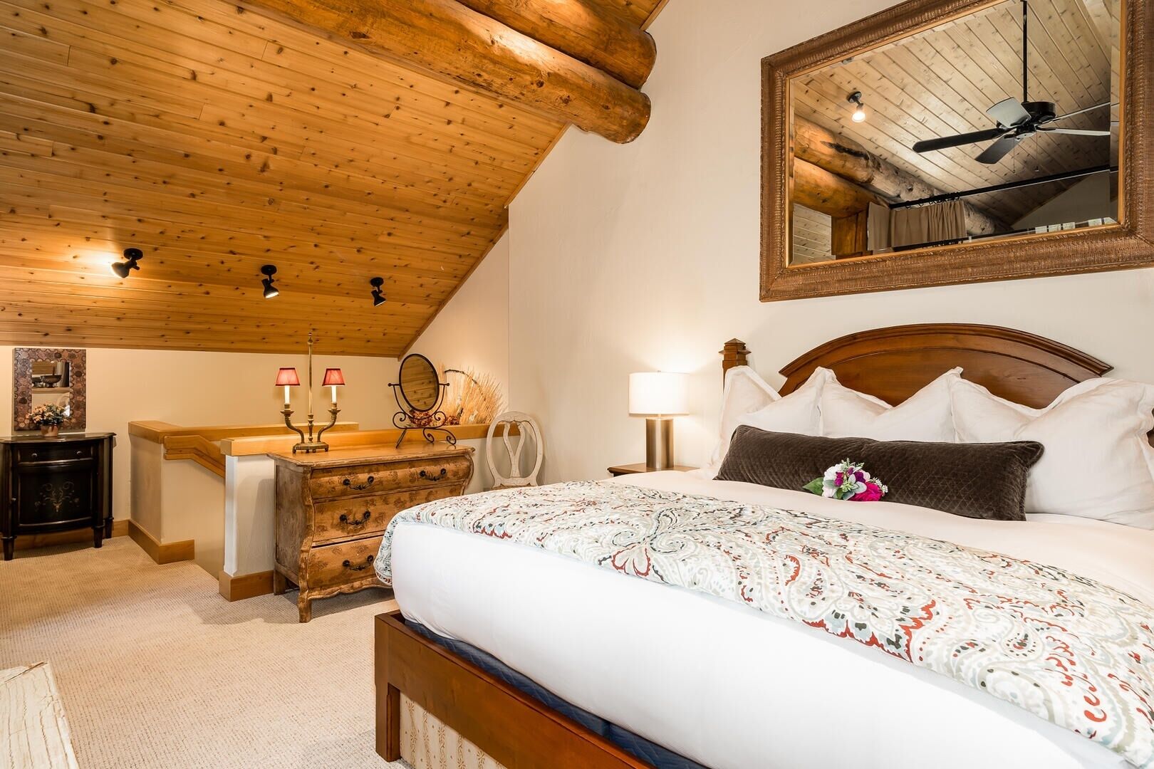 Une chambre avec un plafond en bois, un grand lit recouvert d’une couvre-lit à motifs, une commode, une coiffeuse avec un miroir et un ventilateur de plafond.