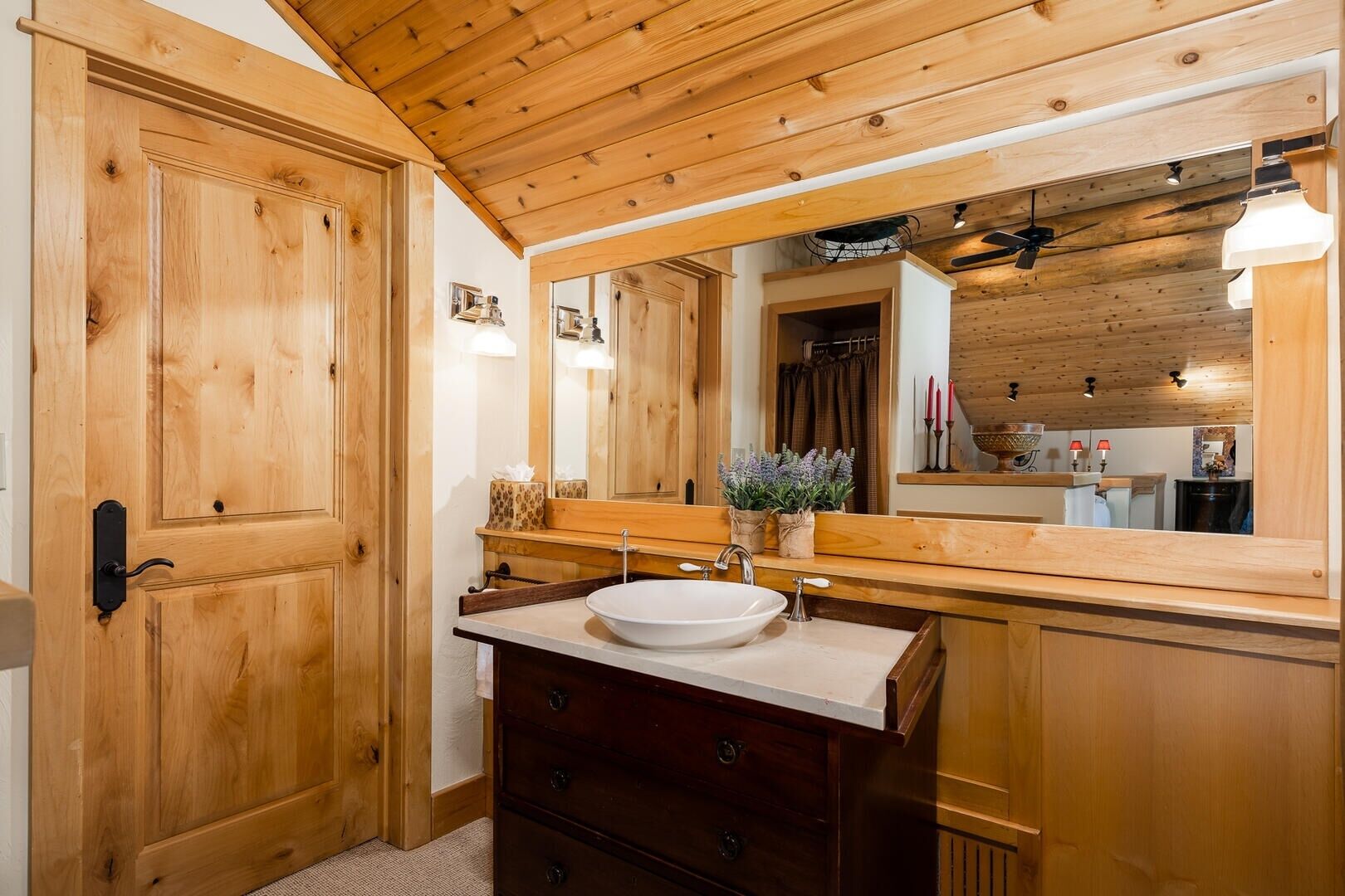 A wooden cabin bathroom with a large mirror, wooden cabinets, a white sink, and a ceiling fan.