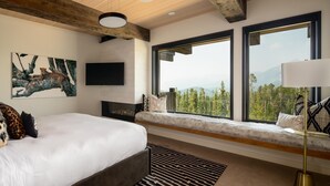 A bedroom with a large bed, a mounted TV, a fireplace, and a window with a mountain view.
