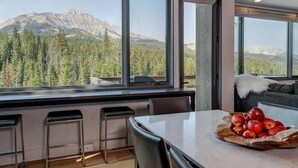 A modern kitchen with large windows offering a mountain view, a dining table with a fruit bowl, and a cozy seating area with a white fur throw. - 17 Lake Cabin 3 Bedroom Home by Moonlight Basin Lodging (Big Sky)