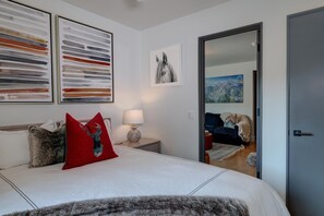 A modern bedroom with a large bed, two abstract paintings, a framed photograph, and a view into another room.