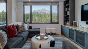A modern living room with a large window offering a mountain view, a dark grey sofa, a coffee table, and a bookshelf with books and decorative items. - 17 Lake Cabin 3 Bedroom Home by Moonlight Basin Lodging (Big Sky)