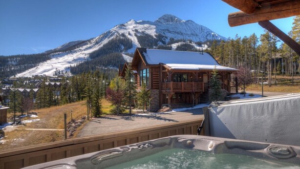 A wooden lodge with a hot tub in front, surrounded by snow-covered mountains and pine trees.