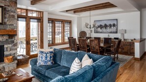 A cozy living room with a stone fireplace, a blue sofa, wooden dining table, and a large window overlooking a snowy landscape.