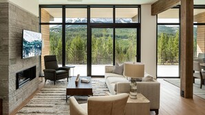 A modern living room with a fireplace, comfortable seating, a large TV, and a view of a mountainous landscape. - 212 Lakelodge 3 Bedroom Condo by Moonlight Basin Lodging (Big Sky)