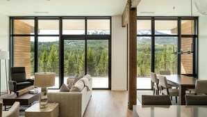 A modern living room with a large window, a sofa, a coffee table, and a dining area. - 212 Lakelodge 3 Bedroom Condo by Moonlight Basin Lodging (Big Sky)